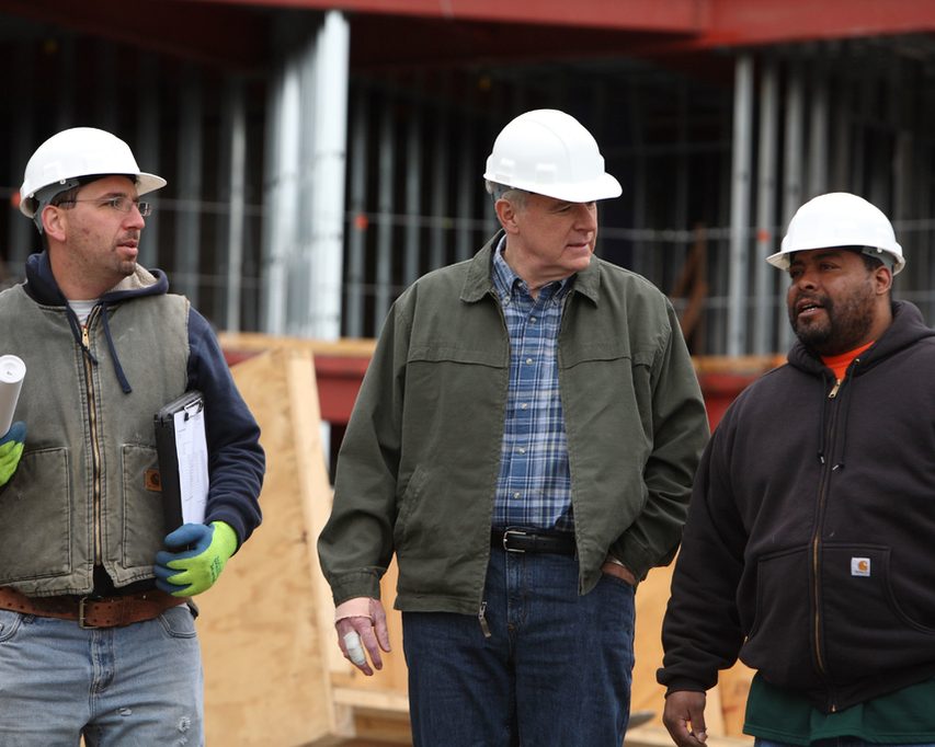 Three members of a building team in hard hats talking on a job site, one holding rolled plans