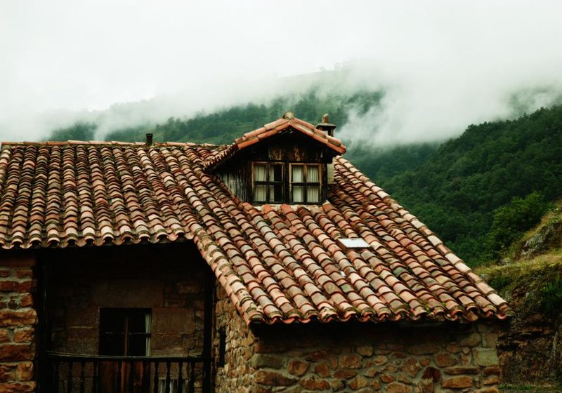 A stone house with a terracotta tile roof and a small dormer window against misty green hills